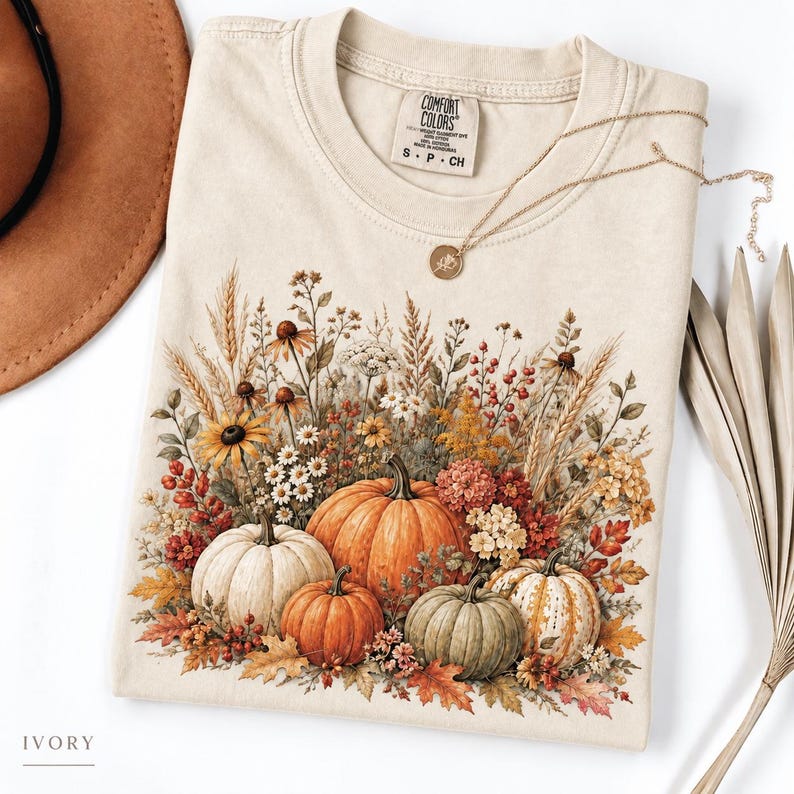 May include: An ivory t-shirt featuring a graphic print of four pumpkins in shades of orange, white, and green, surrounded by dried wheat, wildflowers, and autumn leaves. The shirt has a Comfort Colors label at the neck and a gold-toned necklace.