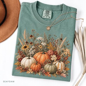 May include: A seafoam green crewneck t-shirt featuring a graphic print of four pumpkins surrounded by dried wheat, wildflowers, and autumn leaves. The inner neck label reads Comfort Colors, S, P, CH. A gold-toned necklace rests on the shirt.