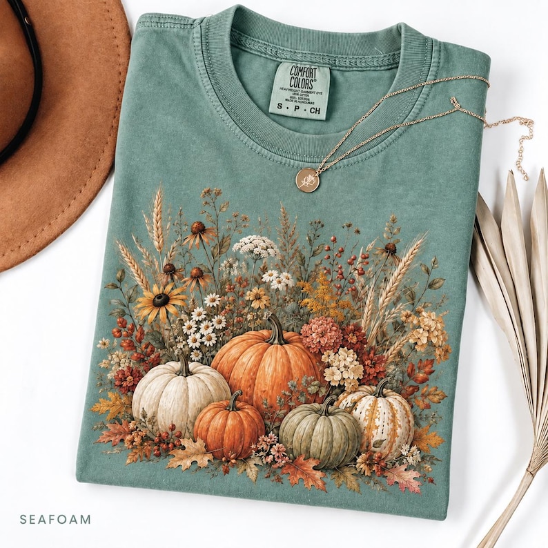 May include: A seafoam green crewneck t-shirt featuring a graphic print of four pumpkins surrounded by dried wheat, wildflowers, and autumn leaves. The inner neck label reads Comfort Colors, S, P, CH. A gold-toned necklace rests on the shirt.