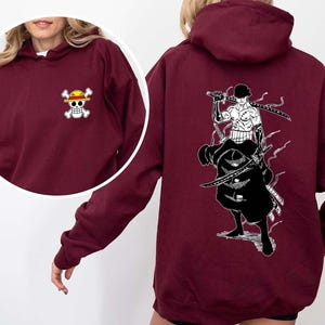K&ouml;nnte beinhalten: Burgunderfarbener Kapuzenpullover mit einem kleinen Totenkopf- und gekreuzten Knochen-Logo auf der Vorderseite und einer gro&szlig;en Schwarz-Wei&szlig;-Illustration eines Schwertk&auml;mpfers auf der R&uuml;ckseite. Der Hoodie hat eine Kapuze und lange &Auml;rmel.