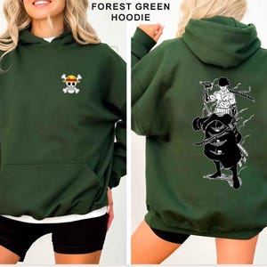K&ouml;nnte beinhalten: Waldgr&uuml;ner Kapuzenpullover mit Fronttasche und Kapuze. Auf der Vorderseite befindet sich ein kleines, farbenfrohes Totenkopf- und gekreuztes Knochen-Design. Auf der R&uuml;ckseite ist eine schwarz-wei&szlig;e Grafik eines Charakters mit Schwertern abgebildet. Der Text "FOREST GREEN HOODIE" steht oben.
