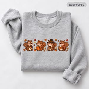 May include: A sport grey crewneck sweatshirt featuring a graphic print of four brown squirrels. The squirrels are depicted with autumn elements including orange pumpkins, brown acorns, a black pilgrim hat, a plaid scarf, and scattered fall leaves.