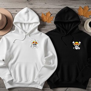 K&ouml;nnte beinhalten: Zwei Kapuzenpullover, einer wei&szlig; und einer schwarz, mit einem Piratensch&auml;del- und Knochenlogo mit Strohhut. Die Kapuzenpullover haben eine Vordertasche und eine Kapuze mit Kordelzug. Ein beiger Hut und eine Tasse Kaffee sind ebenfalls im Bild.
