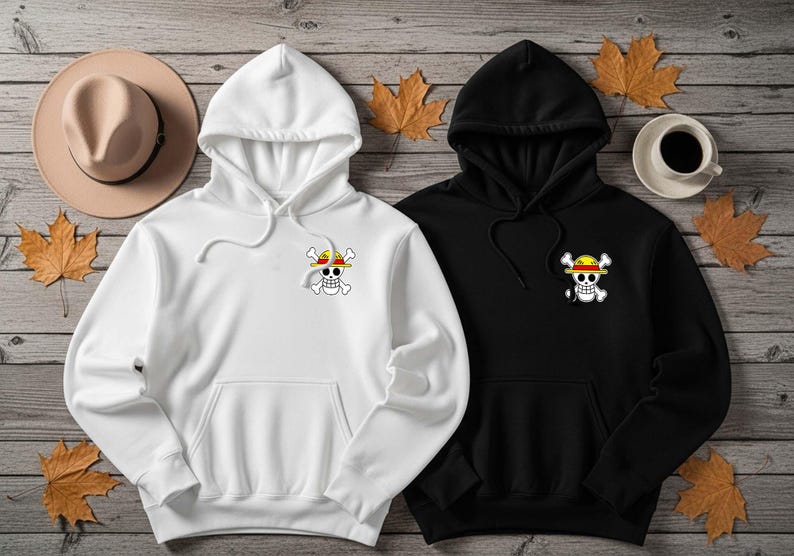 K&ouml;nnte beinhalten: Zwei Kapuzenpullover, einer wei&szlig; und einer schwarz, mit einem Piratensch&auml;del- und Knochenlogo mit Strohhut. Die Kapuzenpullover haben eine Vordertasche und eine Kapuze mit Kordelzug. Ein beiger Hut und eine Tasse Kaffee sind ebenfalls im Bild.