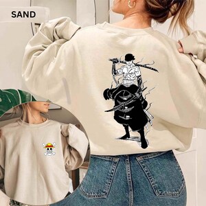 K&ouml;nnte beinhalten: Sandfarbenes Sweatshirt mit einer Grafik eines Charakters mit Schwertern auf dem R&uuml;cken und einem kleinen Emblem auf der Vorderseite. Der Text "SAND" steht oben. Das Sweatshirt ist aus weichem Material.