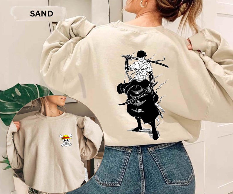 K&ouml;nnte beinhalten: Sandfarbenes Sweatshirt mit einer Grafik eines Charakters mit Schwertern auf dem R&uuml;cken und einem kleinen Emblem auf der Vorderseite. Der Text "SAND" steht oben. Das Sweatshirt ist aus weichem Material.