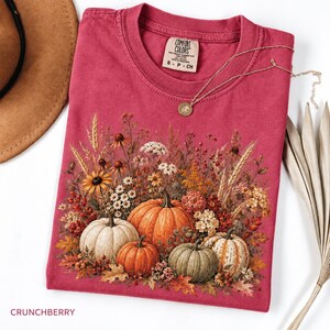 May include: A dark red crewneck t-shirt featuring a graphic print of various pumpkins and autumn wildflowers. The shirt has a Comfort Colors brand label at the inner neckline. A thin gold-toned chain necklace rests on the fabric near the collar.
