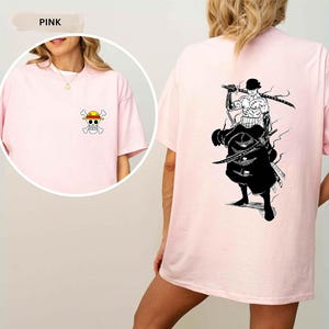 K&ouml;nnte beinhalten: Hellrosa T-Shirt mit einem kleinen Cartoon-Totenkopf und gekreuzten Knochen mit Strohhut auf der Vorderseite. Die R&uuml;ckseite des T-Shirts zeigt eine Schwarz-Wei&szlig;-Illustration einer Figur mit Schwertern.
