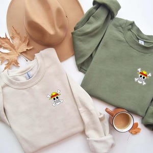 K&ouml;nnte beinhalten: Zwei Sweatshirts mit Rundhalsausschnitt, eines in Creme und eines in Olivgr&uuml;n, jeweils mit einer kleinen, farbenfrohen Totenkopf- und gekreuzten Knochen-Grafik mit gelbem Hut. Ein beiger Hut und ein Herbstblatt sind im Hintergrund.