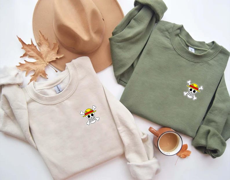 K&ouml;nnte beinhalten: Zwei Sweatshirts mit Rundhalsausschnitt, eines in Creme und eines in Olivgr&uuml;n, jeweils mit einer kleinen, farbenfrohen Totenkopf- und gekreuzten Knochen-Grafik mit gelbem Hut. Ein beiger Hut und ein Herbstblatt sind im Hintergrund.