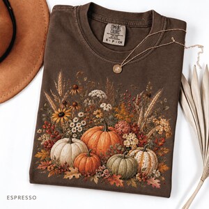 May include: A dark brown short-sleeve t-shirt featuring a graphic print of various pumpkins, wheat stalks, and wildflowers in autumn colours. The inner neck label displays the text Comfort Colors, size S, and indicates the garment is made in Honduras.
