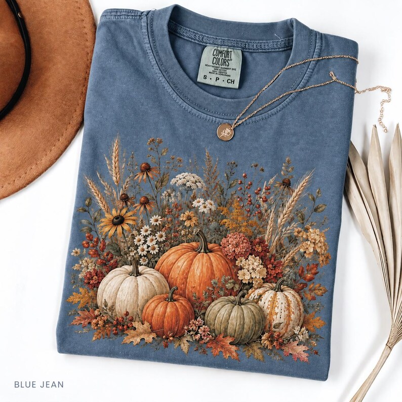 May include: A blue crew neck t-shirt with a Comfort Colors label at the collar. The front features a graphic print of four pumpkins in shades of orange, white, and green, surrounded by dried wheat, wildflowers, and autumn leaves.