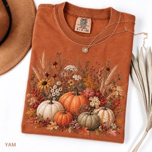 May include: A rust-coloured crew neck t-shirt featuring a graphic print of four pumpkins surrounded by wheat, wildflowers, and autumn leaves. The shirt has a Comfort Colors brand label at the inner neckline and a delicate gold-toned chain necklace.