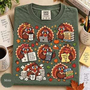 May include: A moss green t-shirt featuring a graphic print of eight cartoon turkeys in various office-themed activities, including using a laptop, holding a coffee cup labeled 'Fueled by Gratitude', writing in a planner, and stacking books.