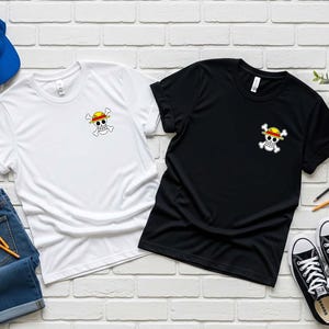 K&ouml;nnte beinhalten: Zwei T-Shirts, eines wei&szlig; und eines schwarz, jeweils mit einer kleinen Grafik eines Sch&auml;dels und gekreuzten Knochen mit einem Strohhut. Die Shirts sind mit einer blauen Baseballkappe, Jeans, Bleistiften und schwarz-wei&szlig;en Turnschuhen ausgelegt.
