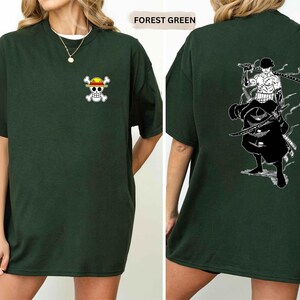 K&ouml;nnte beinhalten: Forstgr&uuml;nes T-Shirt mit einem kleinen Cartoon-Sch&auml;del- und gekreuzten Knochen-Logo auf der Vorderseite. Die R&uuml;ckseite des T-Shirts zeigt eine schwarz-wei&szlig;e Grafik einer Figur mit Schwertern. Das T-Shirt hat eine lockere Passform.