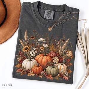 May include: A charcoal grey short-sleeve t-shirt featuring a graphic print of various pumpkins and autumn wildflowers. The shirt has a Comfort Colors label at the neck and is paired with a gold-tone necklace, a brown hat, and dried palm leaves.