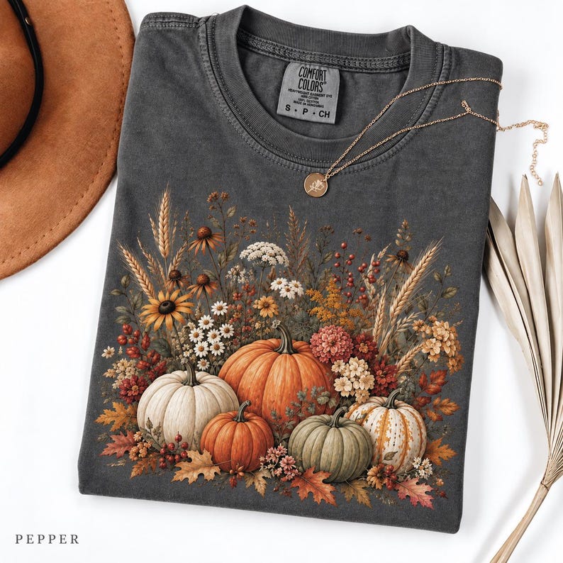 May include: A charcoal grey short-sleeve t-shirt featuring a graphic print of various pumpkins and autumn wildflowers. The shirt has a Comfort Colors label at the neck and is paired with a gold-tone necklace, a brown hat, and dried palm leaves.