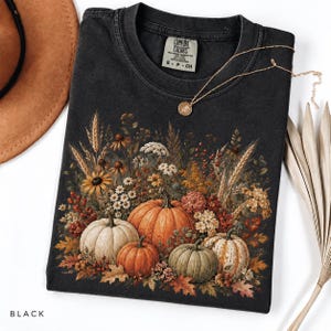 May include: A black t-shirt featuring a graphic print of various pumpkins and autumn wildflowers, including wheat stalks, berries, and fall leaves. The shirt has a Comfort Colors brand label at the neck and a crew neckline with short sleeves.