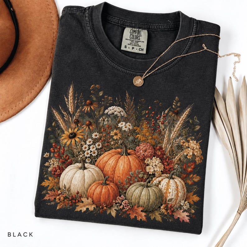 May include: A black t-shirt featuring a graphic print of various pumpkins and autumn wildflowers, including wheat stalks, berries, and fall leaves. The shirt has a Comfort Colors brand label at the neck and a crew neckline with short sleeves.