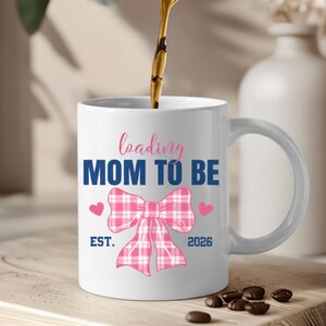 May include: White ceramic mug with the text "loading MOM TO BE" in pink and blue. A pink plaid bow and two pink hearts are featured below the text. The mug also includes "EST. 2026". Coffee is being poured into the mug.