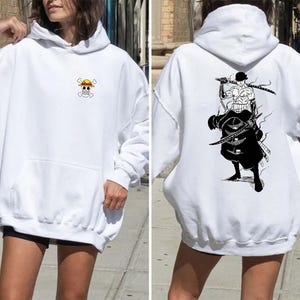 K&ouml;nnte beinhalten: Wei&szlig;er Kapuzenpullover mit Fronttasche und Kapuze. Auf der Vorderseite befindet sich ein kleines, farbenfrohes Piratenflaggen-Emblem. Auf der R&uuml;ckseite ist eine Schwarz-Wei&szlig;-Illustration eines Schwertk&auml;mpfers mit drei Schwertern abgebildet.