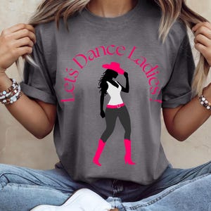 Peut inclure: T-shirt gris avec le texte rose "Let's Dance Ladies!". Le t-shirt présente une silhouette de femme en haut blanc, pantalon gris, bottes roses et chapeau rose. Le motif est centré sur le t-shirt.