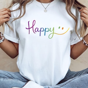 Könnte beinhalten: Weißes T-Shirt mit dem Wort "Happy" in einem Regenbogen-Farbverlauf und einem Smiley. Das Shirt ist ein lässiger Stil, geeignet für den Alltag. Das Design ist einfach und fröhlich.