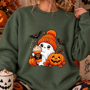Könnte beinhalten: Ein olivgrüner Rundhals-Pullover mit einer Grafik eines weißen Geistes, der eine orangefarbene Mütze trägt und einen Becher mit der Aufschrift PUMPKIN SPICE hält. Das Design zeigt zwei schwarze Fledermäuse, zwei orange Kürbisse und Herbstblätter.