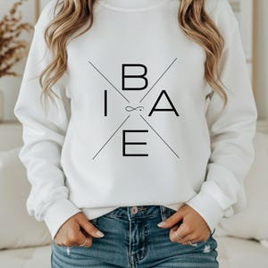 May include: White sweatshirt with the word "B.I.A.E." in black, arranged in a cross pattern. The sweatshirt has long sleeves and a crew neck. The model is wearing blue jeans.