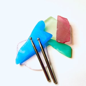 Puede incluir: Dos brocas de metal plateado con punta esférica se colocan sobre una colección de piezas de vidrio de colores de forma irregular. Las piezas de vidrio son en tonos de azul, verde, rosa y blanco, creando una composición vibrante.