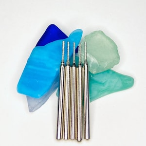 Può includere: Cinque punte diamantate argentate per l'uso con uno strumento rotativo. Le punte sono disposte in fila su uno sfondo di vetro blu e verde.