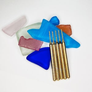 Puede incluir: Una variedad de piezas de vidrio de mar coloridas en tonos rosa, azul, verde y naranja. Cinco herramientas doradas y delgadas con puntas texturizadas están dispuestas sobre un fondo blanco. El vidrio de mar añade un toque de belleza natural.