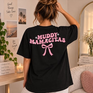 Könnte beinhalten: Ein schwarzes Kurzarm-T-Shirt mit dem rosa Schriftzug "MUDDY MAMACITAS" auf der Rückseite in wellenförmiger Schrift, ergänzt durch kleine vierzackige Sternsymbole und eine rosa Schleifengrafik unter dem Text.