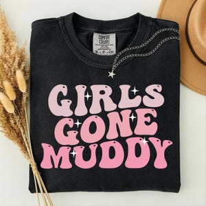 May include: Black t-shirt with the text "GIRLS GONE MUDDY" in pink and light pink bubble letters. A silver necklace with a star pendant is draped on the shirt. A brown hat is visible in the background.