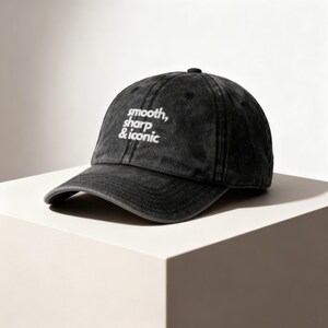 Könnte beinhalten: Schwarze Baseballkappe mit den in Weiß gestickten Worten "smooth, sharp & iconic". Die Kappe hat einen gebogenen Schirm und einen Knopf oben. Der Hut ist aus einem abgenutzten, verblichenen Material.
