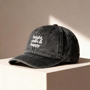 Könnte beinhalten: Eine schwarze Baseballkappe mit dem weißen Schriftzug "bright, calm & happy". Die Kappe hat einen gebogenen Schirm und einen Knopf oben. Der Hut ist aus weichem, abgenutztem Material.