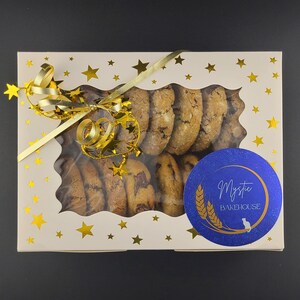 May include: A box of cookies with a clear window, decorated with gold stars and a gold ribbon. The cookies are various shades of brown and have chocolate chips. A blue sticker with the text "Mystic Bakehouse" is on the box.