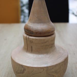 Peut inclure: Un pot en bois artisanal avec un couvercle conique. Le pot a une base arrondie, une section médiane cylindrique et un couvercle qui se rétrécit en pointe. Le bois est de couleur claire et naturelle, avec des veines visibles et quelques nœuds plus foncés.