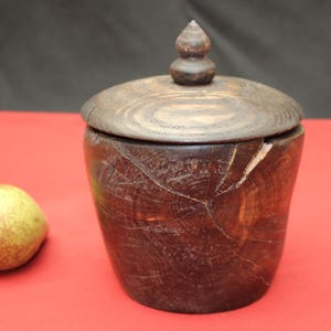 May include: A handcrafted wooden jar with a lid, featuring a dark brown finish and visible wood grain. The jar has a cylindrical shape with a lid and a small knob on top. A pear is next to the jar.
