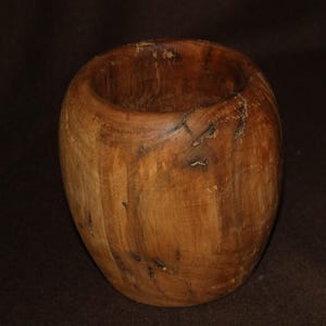 Puede incluir: Un cuenco de madera hecho a mano, de forma redondeada y con una amplia abertura. El cuenco es de madera marrón claro con vetas y nudos más oscuros, mostrando la veta y textura naturales. El interior es liso, y el exterior tiene una textura ligeramente más rugosa.