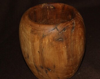 May include: A wooden bowl with a rounded form and a broad opening. The bowl is crafted from light brown wood, featuring darker streaks and knots that highlight the natural grain and texture. The interior is smooth, while the exterior has a slightly rougher texture.