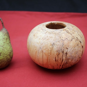 Può includere: Una pera verde con macchie marroni e un vaso sferico in legno con un foro nella parte superiore. Il vaso è di colore marrone chiaro con striature marrone scuro. Entrambi gli oggetti sono su una superficie rossa.