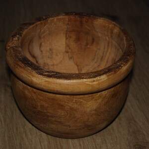 May include: A wooden bowl with a natural finish. The bowl has a rounded shape with a wide rim and a textured surface, showing the wood grain. This decorative bowl is suitable for serving or display, and is a rustic item.
