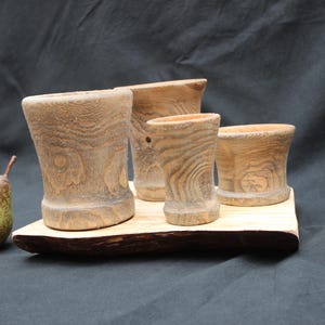 Può includere: Quattro vasi in legno con finitura naturale venata, posti su un vassoio di legno. Una pera verde è appoggiata a sinistra del vassoio. I vasi variano per dimensioni e forma, con un colore marrone chiaro.