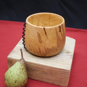 Op de afbeelding: Een handgemaakte houten beker met een natuurlijke afwerking, met een donker spiraalvormig detail aan de zijkant. De beker staat op een houten blok, met een groene peer ernaast. De achtergrond is een rood oppervlak met een zwarte achtergrond.