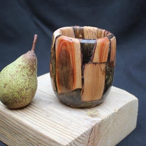 Peut inclure: Une tasse en bois faite à la main avec un motif en mosaïque de différentes teintes de bois, dont des bruns clairs et foncés, et des accents orange-rougeâtre. Une poire verte avec des taches brunes est posée à côté de la tasse sur un bloc de bois. Le fond est sombre.