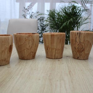 Peut inclure: Quatre gobelets en bois avec une finition marron clair naturelle sont disposés sur une table en bois clair. Les gobelets ont une forme légèrement conique et montrent le grain naturel du bois. L'arrière-plan comprend une fenêtre et de la verdure.