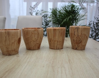 May include: Four wooden cups with a natural, light brown finish are arranged on a light-coloured wooden table. The cups have a slightly tapered shape and show the natural grain of the wood. The background includes a window and greenery.