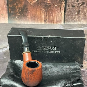 Könnte beinhalten: Eine Tabakpfeife mit einem rotbraunen Kopf und einem schwarzen Mundstück, die auf einem schwarzen Lederbeutel liegt. Die Pfeife wird von einer schwarzen Schachtel mit dem Text "SANDA DURABLE TOBACCO PIPE" begleitet.